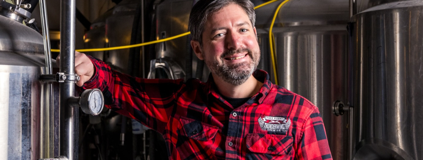 A man in a red plaid shirt stands smiling inside a brewery, resting his arm on a stainless steel brewing tank surrounded by hoses and equipment.