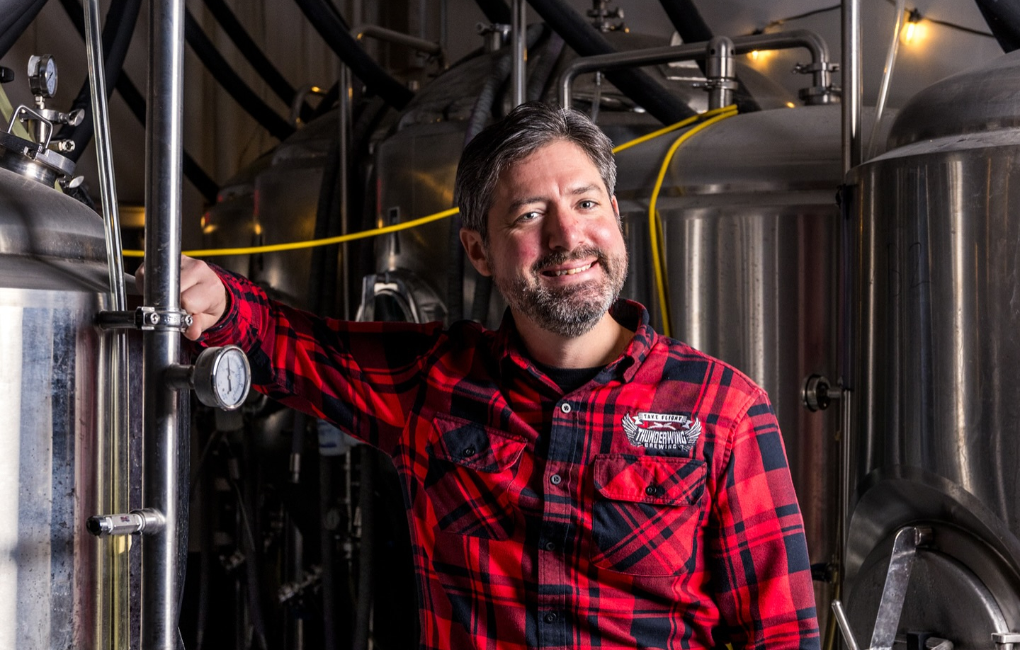 A man in a red plaid shirt stands smiling inside a brewery, resting his arm on a stainless steel brewing tank surrounded by hoses and equipment.