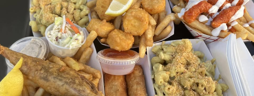 A tray of assorted food truck vegan dishes featuring fried fish, mac and cheese, yucca fries. hush puppies, egg rolls, coleslaw, and dipping sauces, all served in paper food trays.