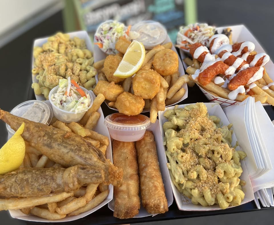 A tray of assorted food truck vegan dishes featuring fried fish, mac and cheese, yucca fries. hush puppies, egg rolls, coleslaw, and dipping sauces, all served in paper food trays.